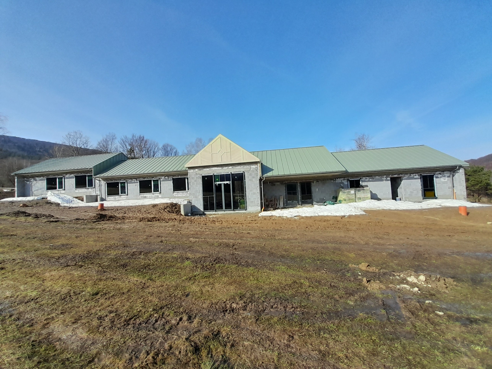 Budynek w stanie surowym zamkniętym z zielonym dachem blachodachówkowym i szarymi ścianami z pustaków, z widocznym oknem i drzwiami, na tle błotnistego terenu i błękitnego nieba.