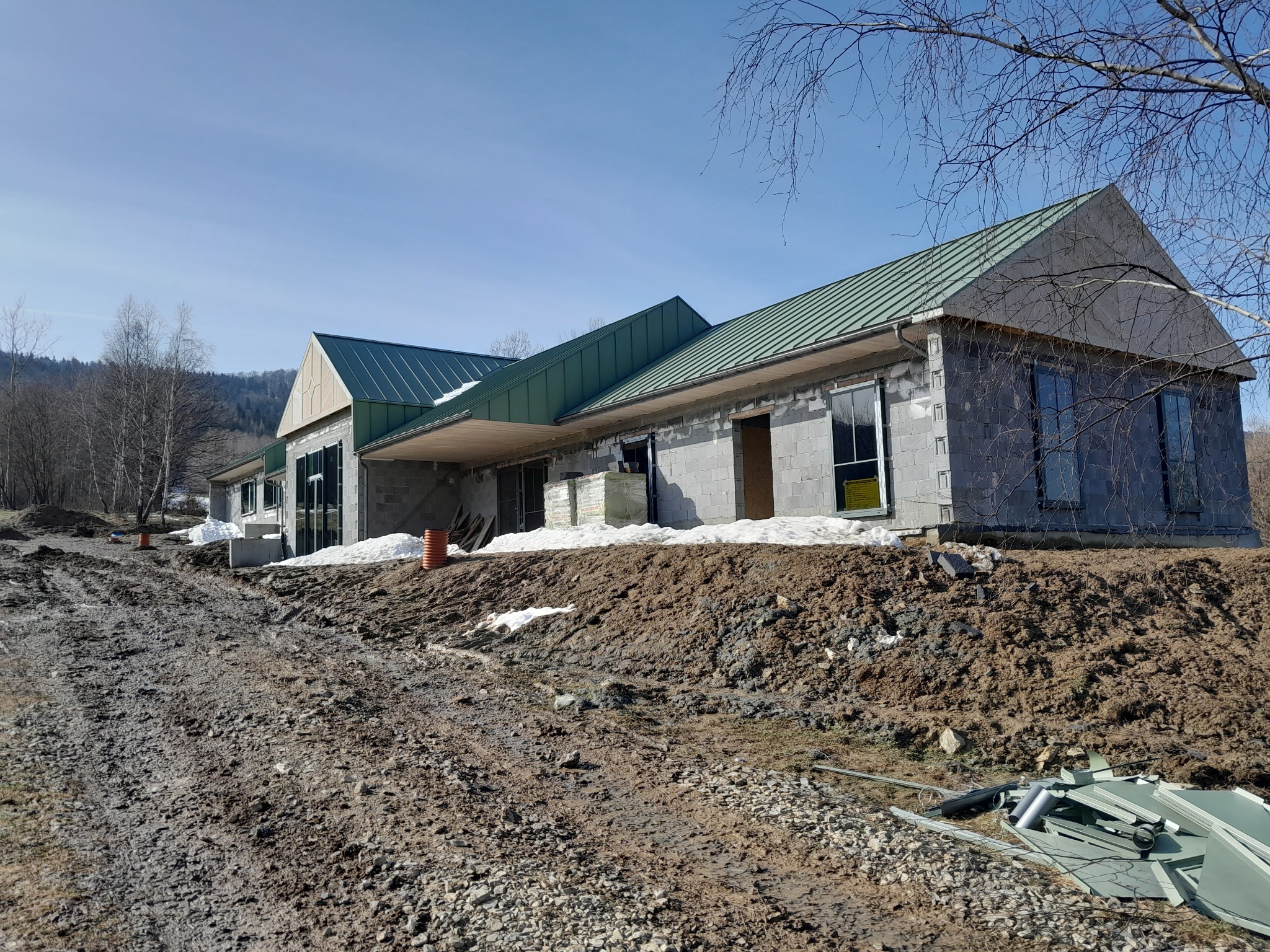 Surowy stan budowy domu jednorodzinnego z zielonym dachem na wsi, widoczne bloczki betonowe, ziemia i resztki śniegu.