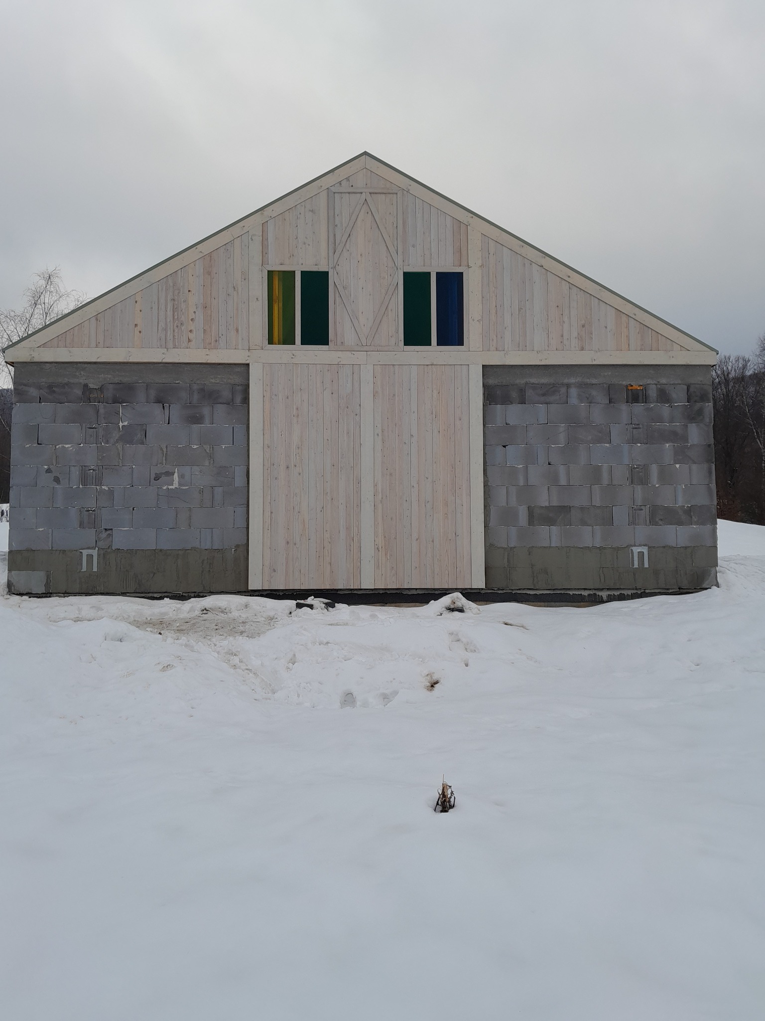 Budynek w trakcie budowy z szarych betonowych bloków i drewnianym szczytem oraz dwiema kolorowymi szybami okiennymi, częściowo zasypany śniegiem.