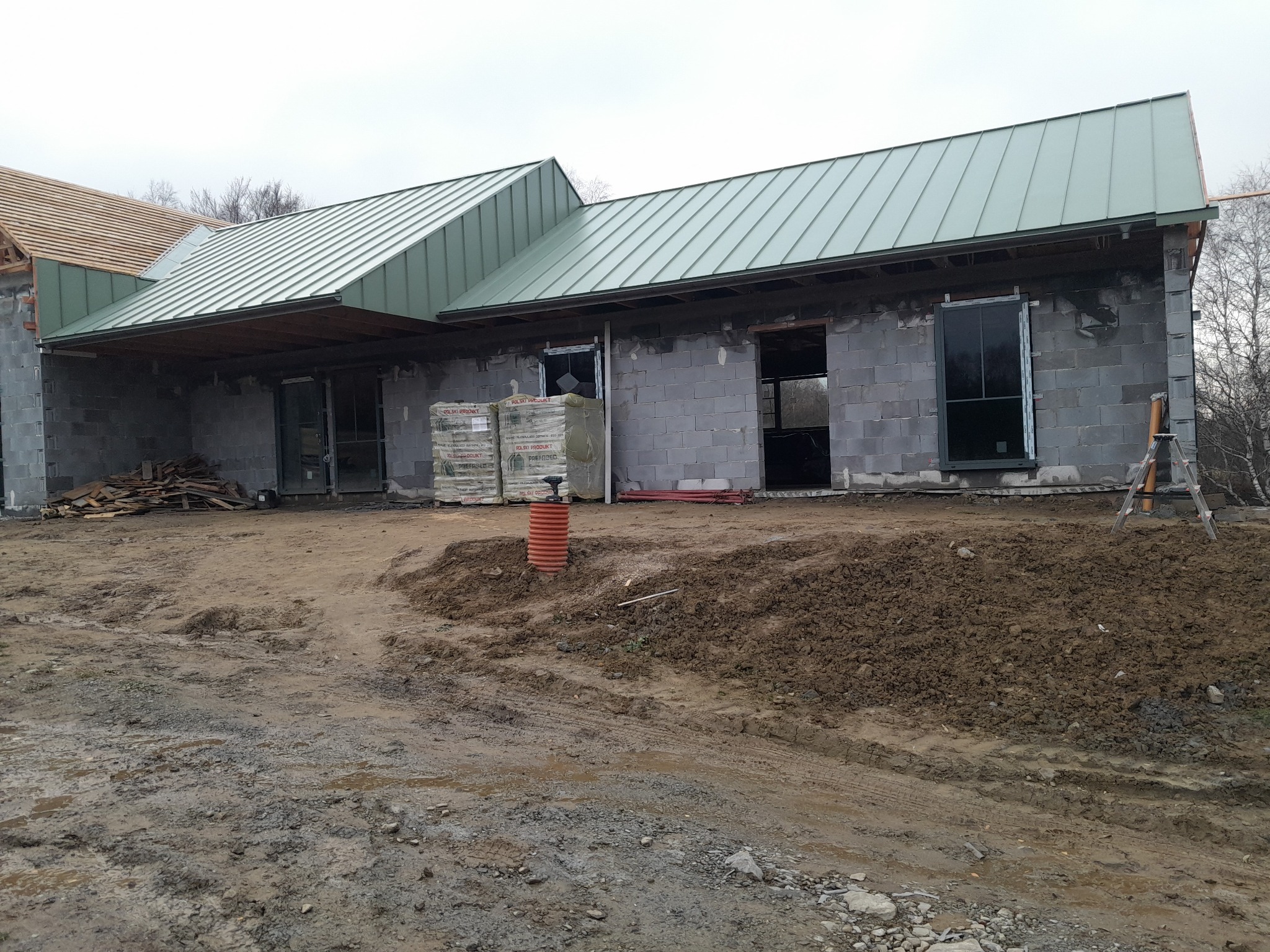Surowy stan budowy domu z szarego bloczka w Grybowie, z widocznym zielonym dachem z blachy trapezowej, stertą materiałów budowlanych i błotnistym terenem wokół.