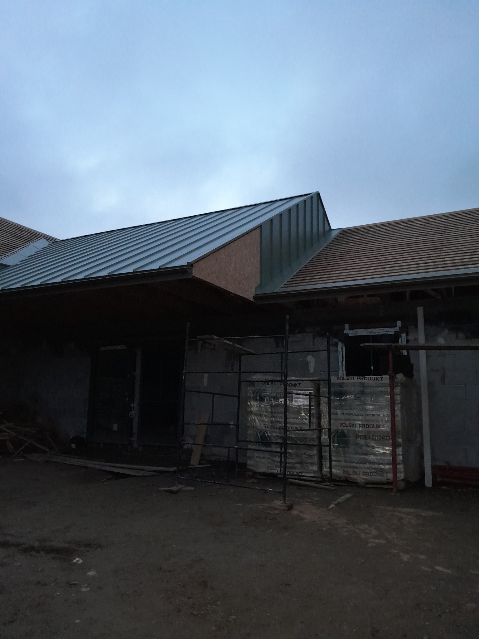 Wznoszona konstrukcja domu z widocznym fragmentem dachu pokrytego blachą trapezową, z rusztowaniem i materiałami budowlanymi na pierwszym planie, sfotografowana w pochmurny dzień.