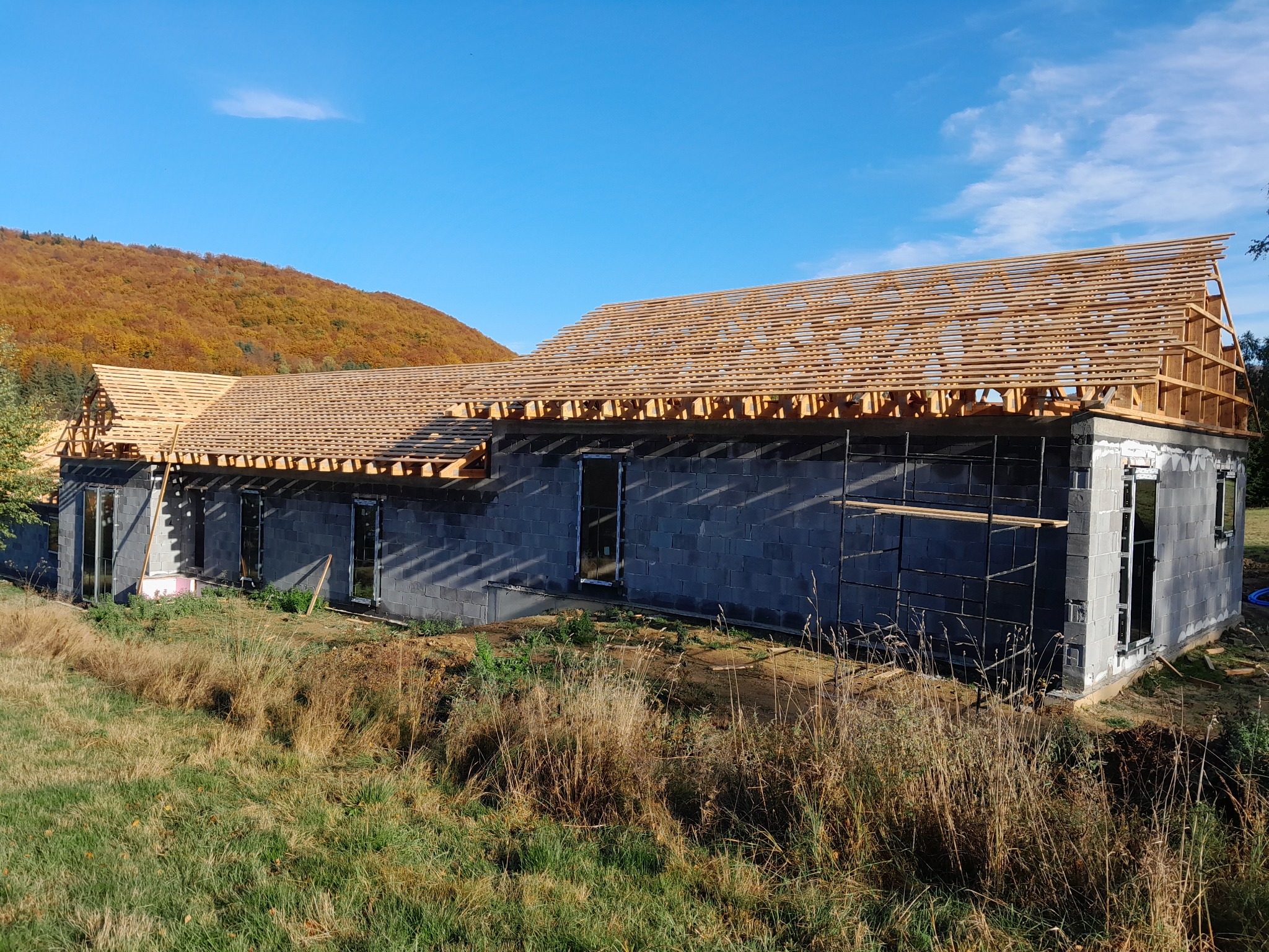 Dom w stanie surowym otwartym z widoczną konstrukcją drewnianą dachu na tle jesiennego krajobrazu z zalesionym wzgórzem.