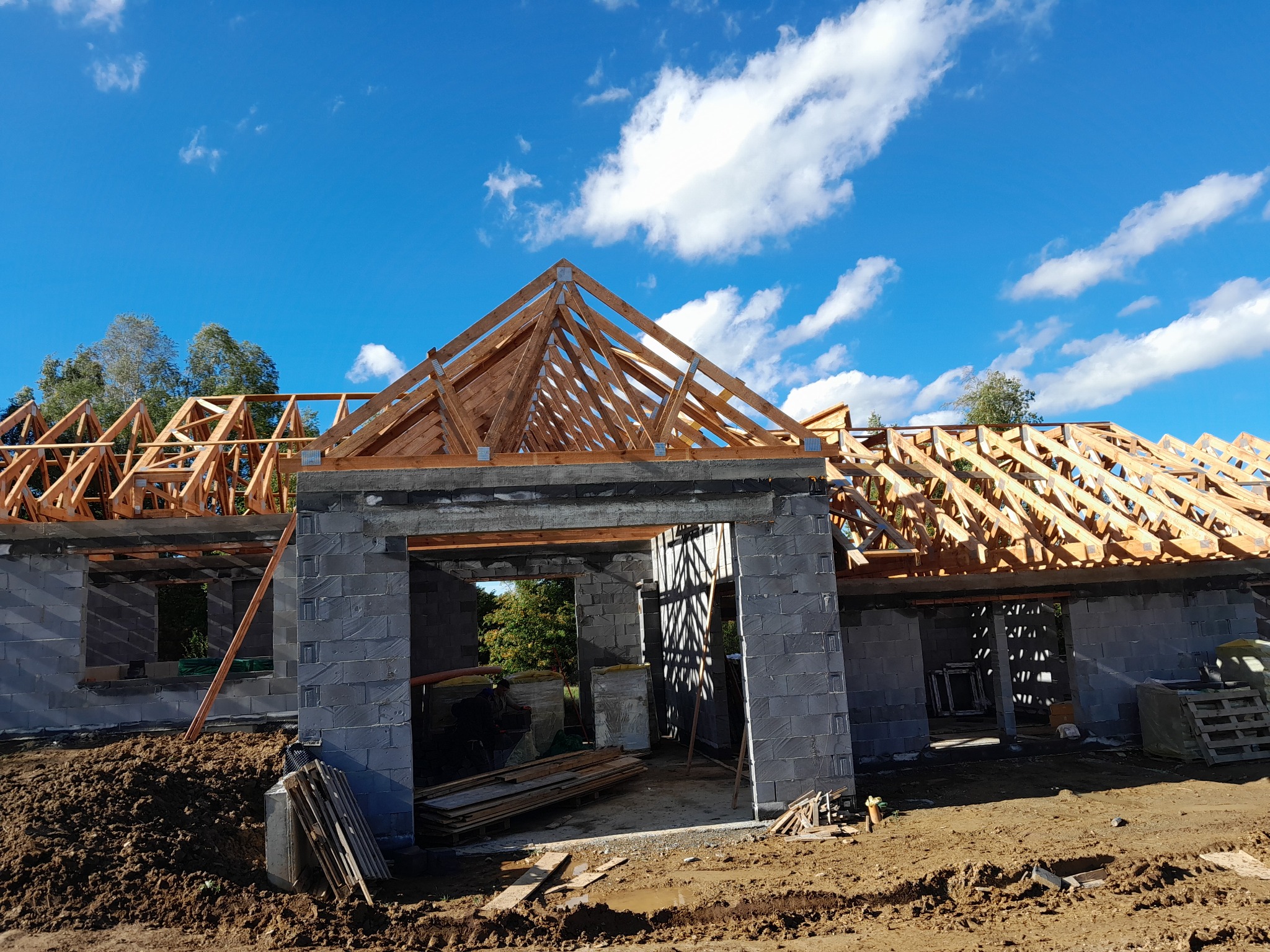 Surowy stan domu w trakcie budowy z szarych bloczków, widoczna drewniana konstrukcja dachu na tle błękitnego nieba z chmurami, teren wokół w stanie budowy.