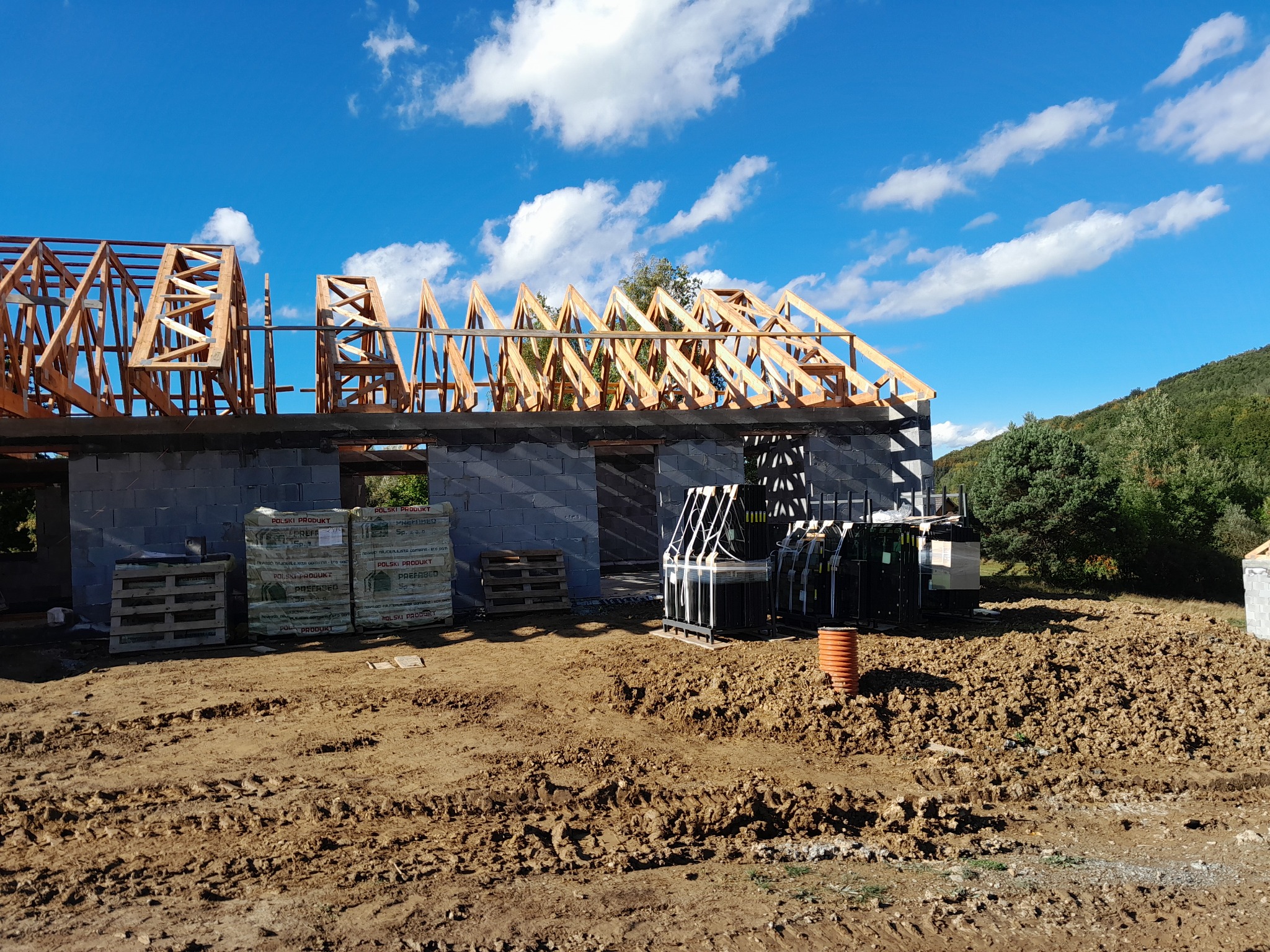 Wznoszona konstrukcja domu z drewnianym szkieletem dachu na murze z bloczków betonowych, palety z materiałami budowlanymi i pakiet okien na placu budowy z ziemią i śladami opon, w tle zielone...