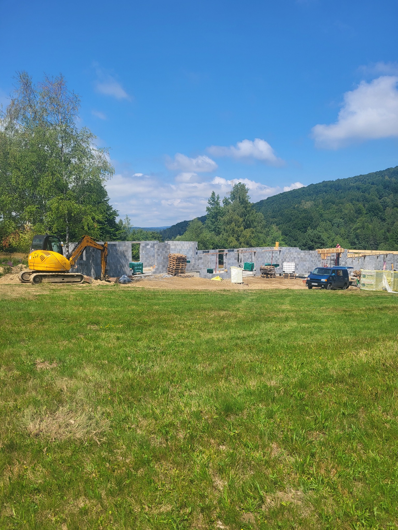 Budowa domu jednorodzinnego w trakcie wznoszenia ścian z betonu komórkowego, widoczny żółty mini-koparka JCB, palety z materiałami budowlanymi i granatowy samochód na tle zielonego trawnika...