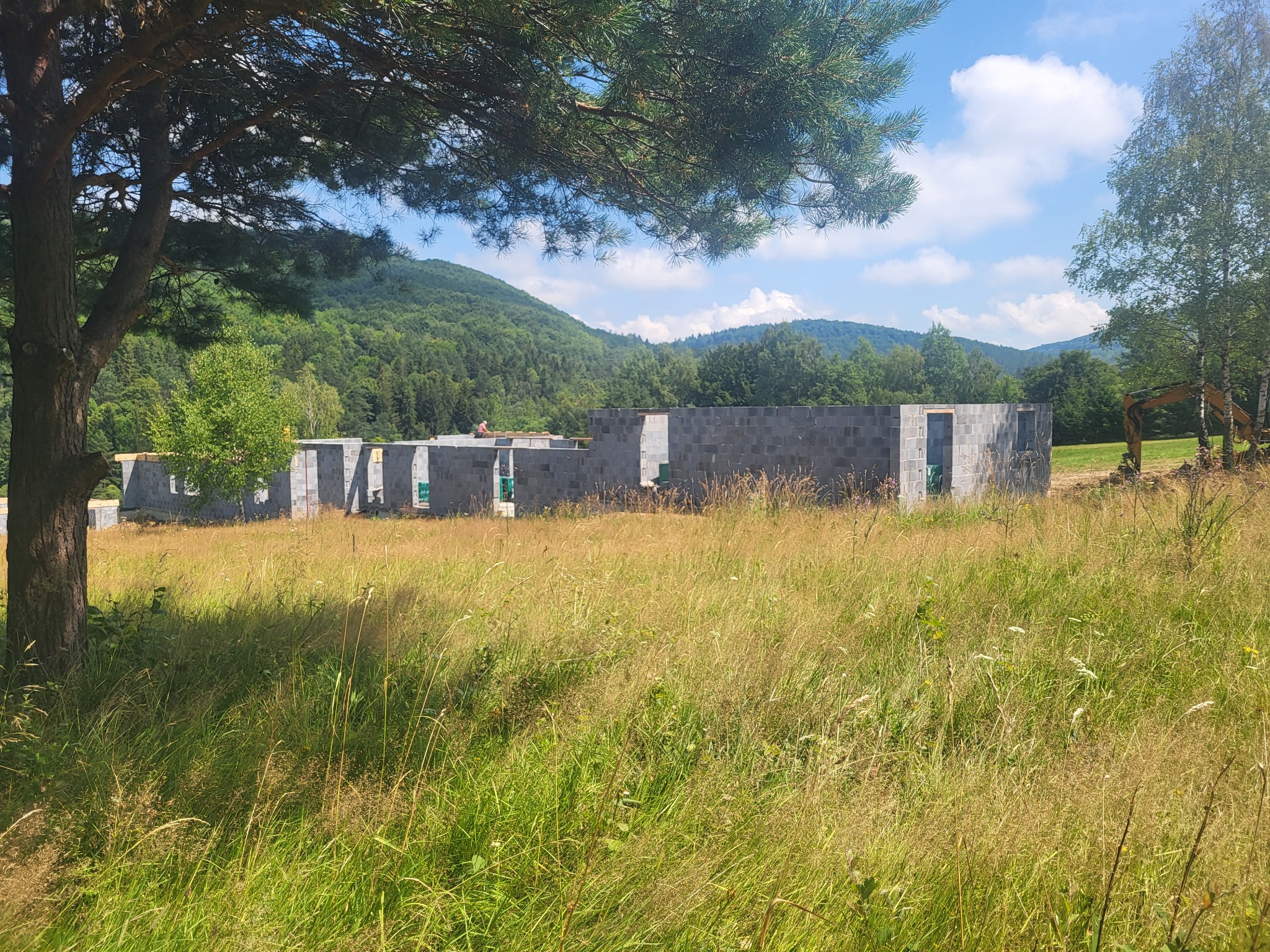 Niedokończony budynek z szarego bloczka na tle łąki i zalesionych wzgórz w słoneczny dzień, widoczny częściowo pod drzewem iglastym, z koparką w tle.