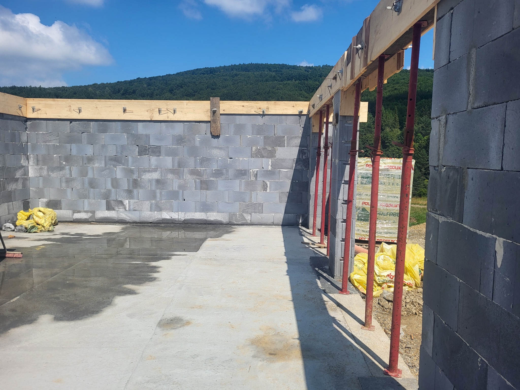 Wnętrze budynku w trakcie budowy z szarych bloczków, częściowo zalana posadzka, drewniane szalunki podtrzymywane przez czerwone stemple, góry w tle.