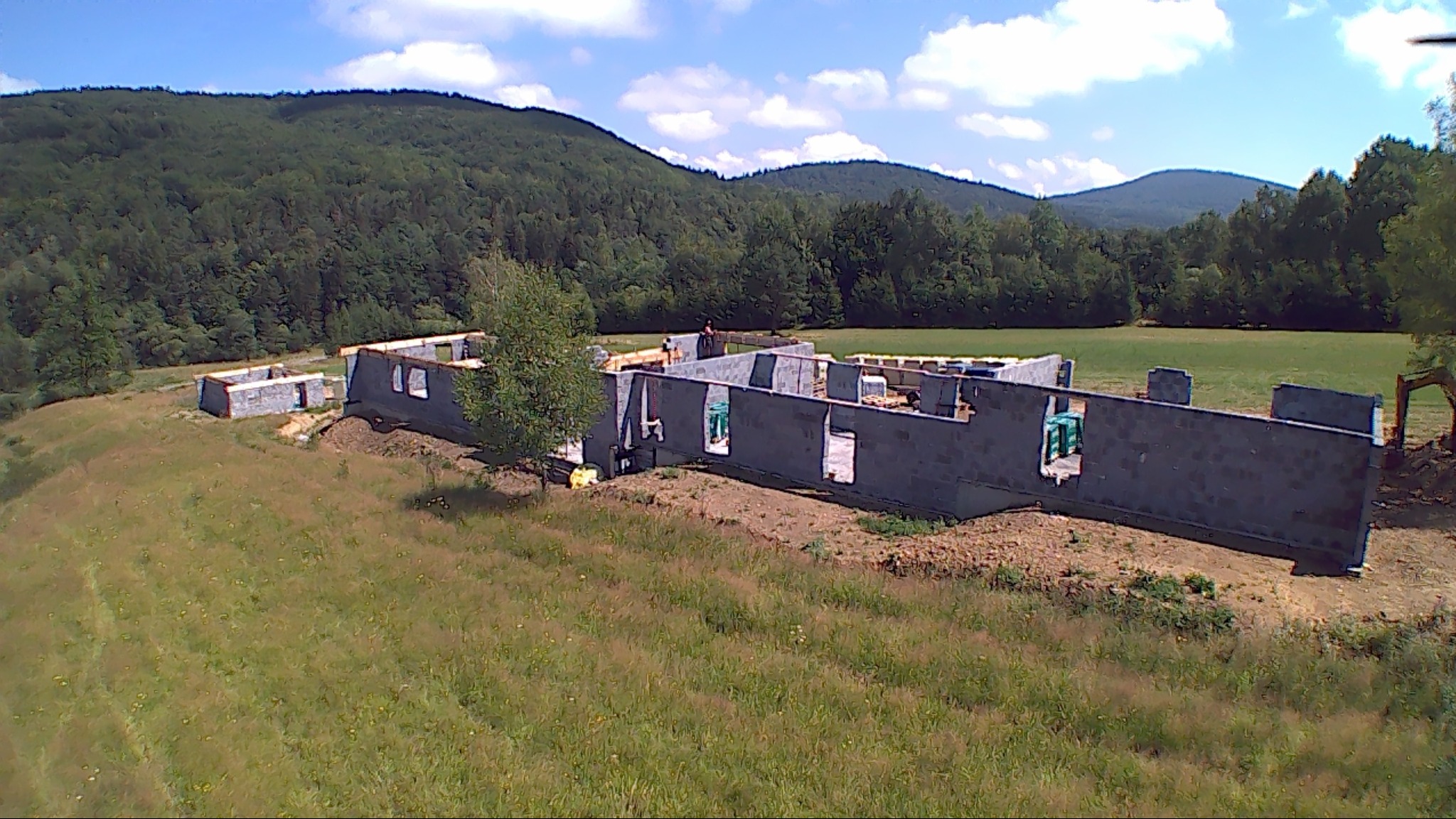 Budowa domu z bloczków betonowych na etapie wznoszenia ścian fundamentowych, widoczny pracownik na tle zielonych wzgórz i błękitnego nieba z chmurami.