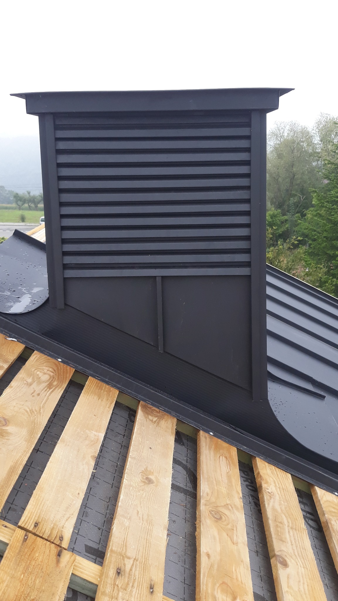 Czarny komin wentylacyjny z poziomymi żaluzjami na dachu z blachy na rąbek stojący. Widoczne drewniane łaty pod blachą. Tło stanowią drzewa i zamazane wzgórza.