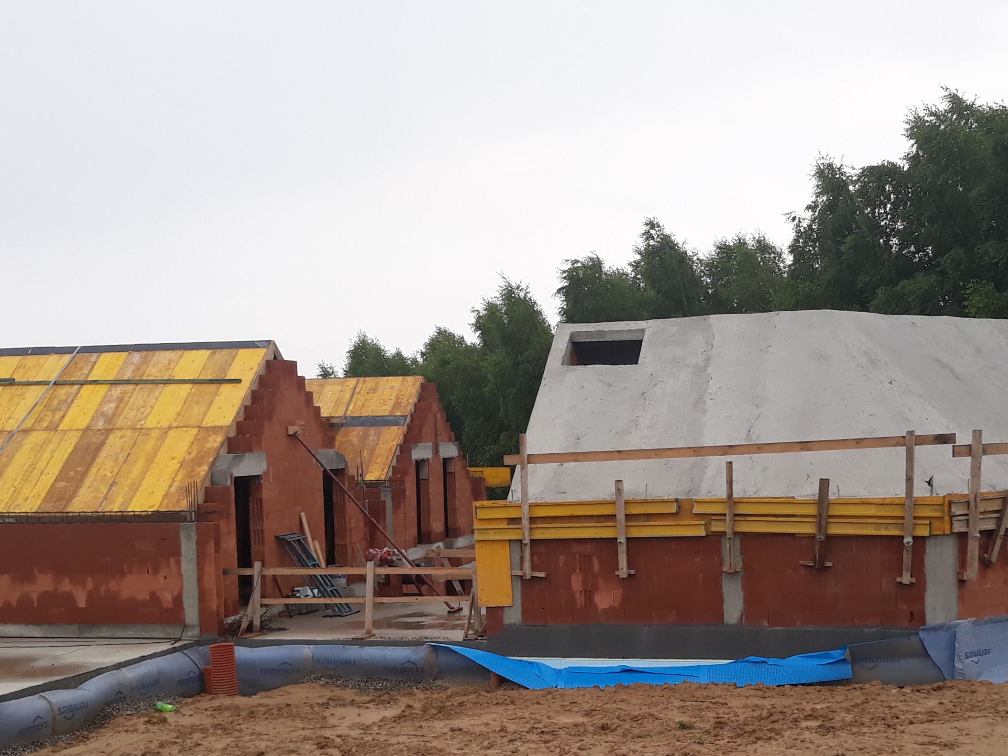 Budowa domów w trakcie: murowane ściany z żółtym szalunkiem na dachu, betonowy element z otworem, zabezpieczone fundamenty, ziemia w tle i częściowo pochmurne niebo.
