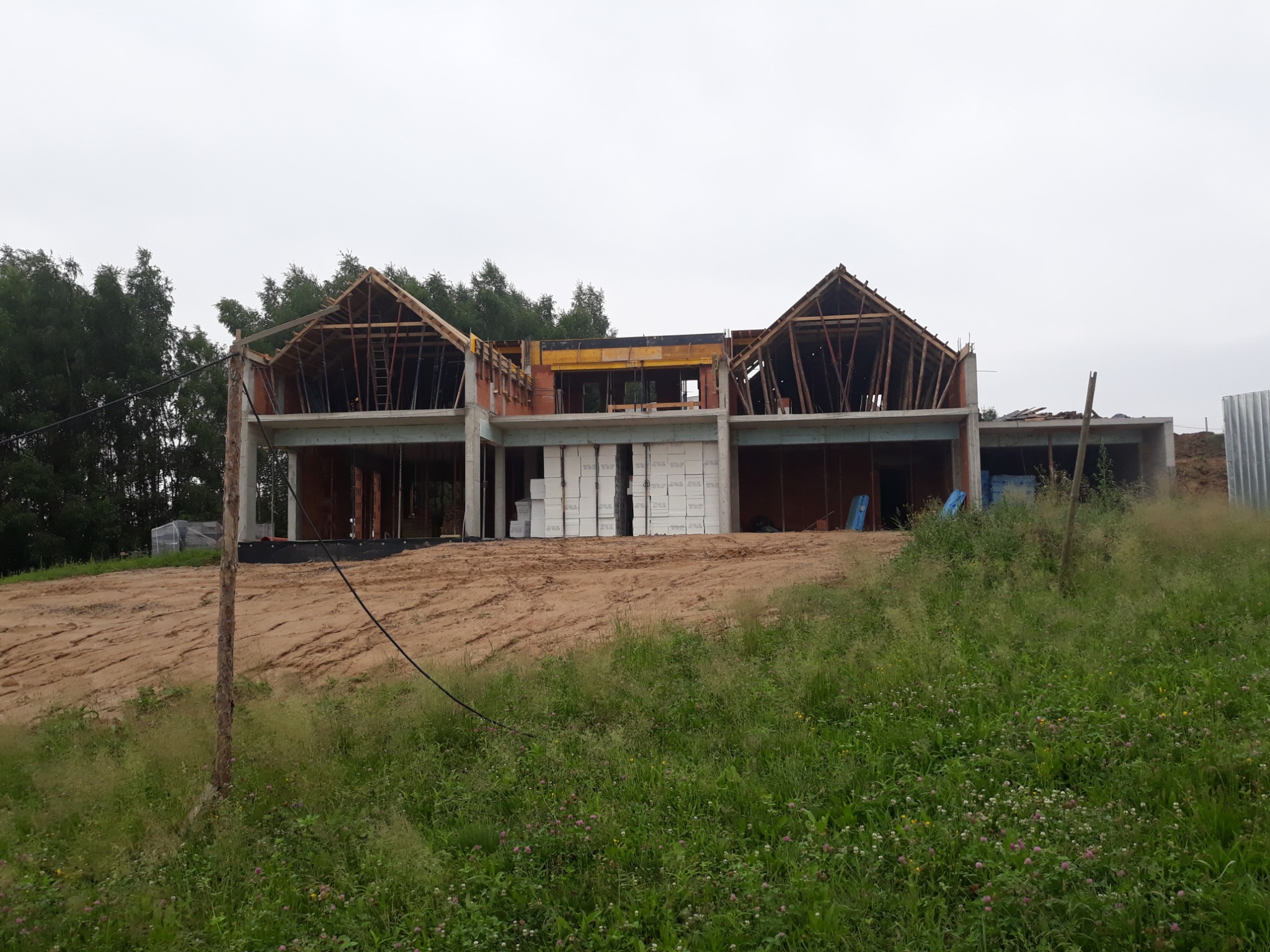 Wznoszenie dwukondygnacyjnego domu z elementami drewnianymi konstrukcji dachu. Widoczne bloczki i betonowe słupy. Teren wokół w stanie surowym, zarośnięty trawą.