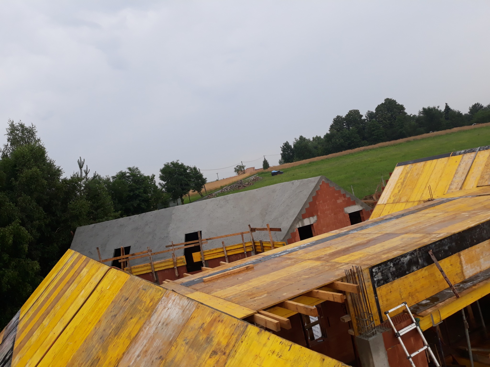 Budowa domu w trakcie realizacji: widok z góry na szalunki stropowe, ściany z pustaków i betonowy dach. W tle zielone wzgórza i pochmurne niebo.