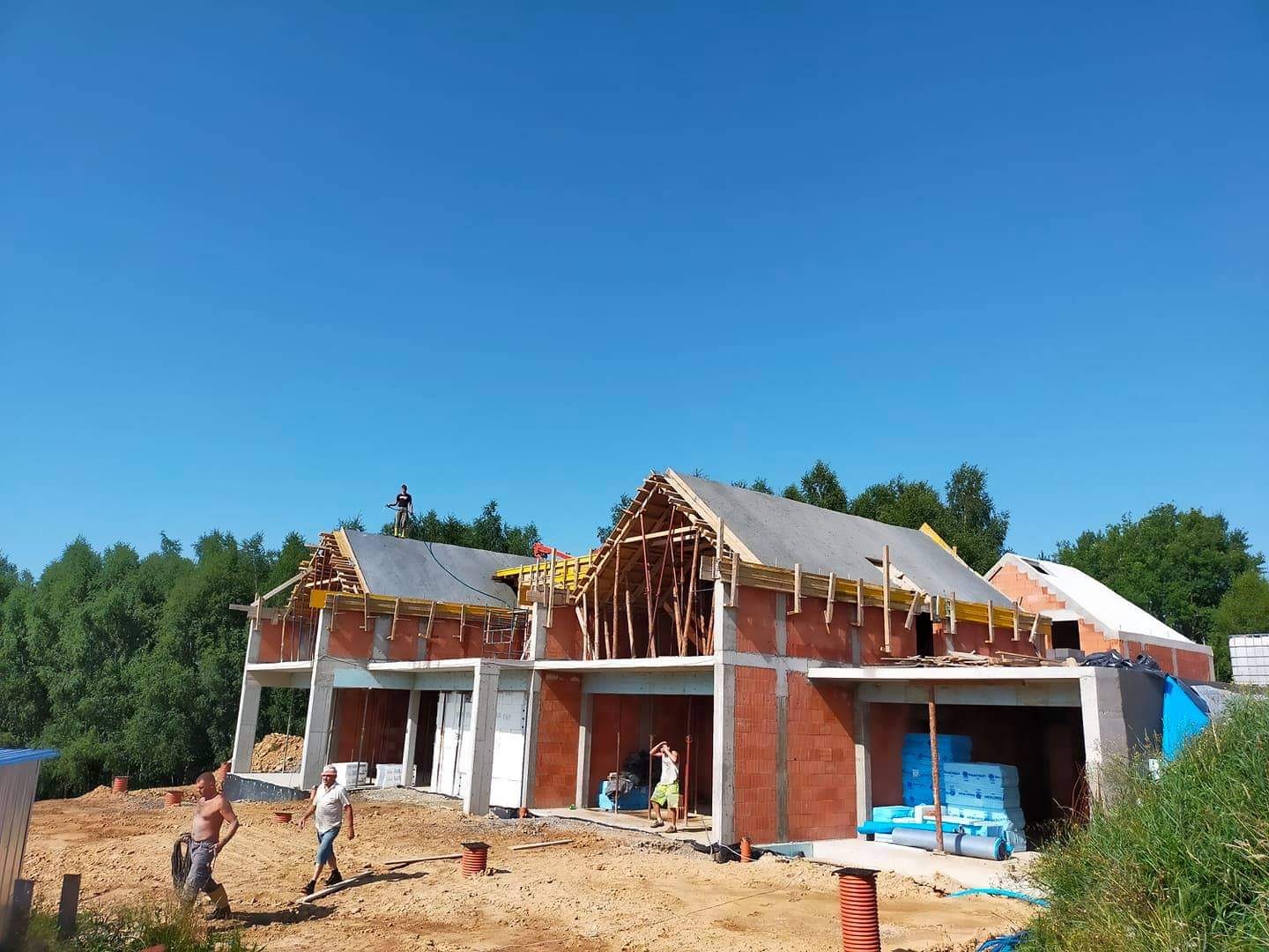 Budowa domu jednorodzinnego w Grybowie: ściany z czerwonej cegły, drewniana konstrukcja dachu w trakcie montażu, widoczni pracownicy budowlani.