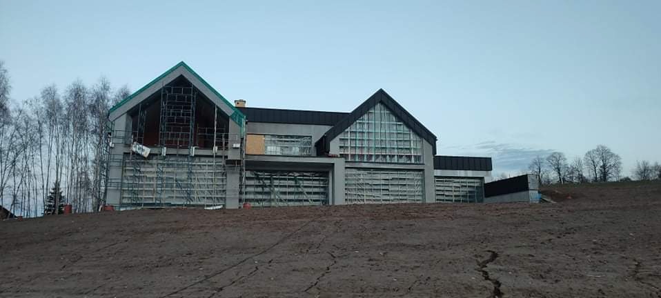 Nowoczesny dom w trakcie budowy, widoczne rusztowania i niezabudowane okna. Surowy krajobraz z ziemią na pierwszym planie. Architektura z elementami szkła i ciemnego dachu.