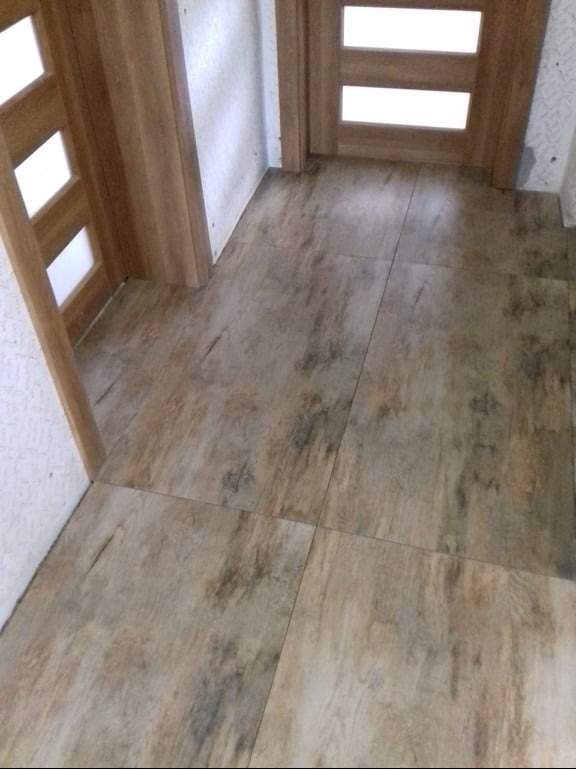 Ułożona podłoga z płytek imitujących drewno w korytarzu, widoczne dwie ościeżnice drzwiowe z drewnianymi drzwiami z przeszkleniami.