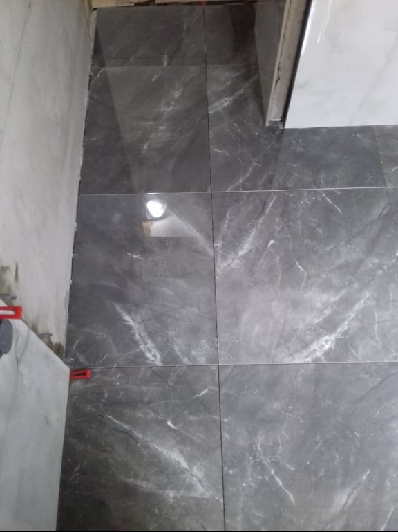 Układanie szarych płytek podłogowych z marmurkowym wzorem, widoczne czerwone kliny poziomujące i fragment surowej ściany.