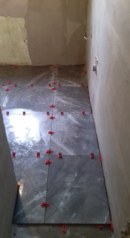 Układanie ciemnoszarych płytek podłogowych z marmurkowym wzorem, z użyciem czerwonych krzyżyków dystansowych w surowym wnętrzu łazienki w trakcie remontu.