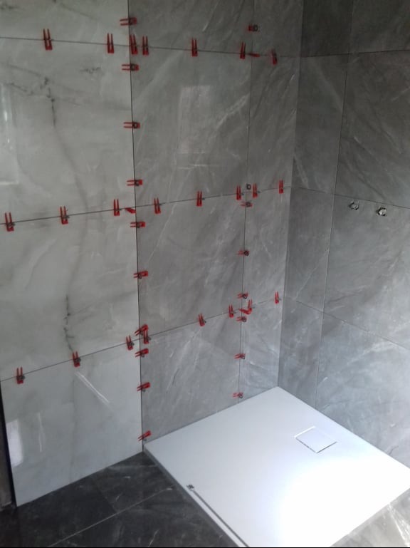 Szare płytki imitujące marmur, ułożone w kabinie prysznicowej z białym brodzikiem, z czerwonymi klipsami poziomującymi i czarnymi krzyżykami dystansującymi. Widoczne niedokończone fugowanie.