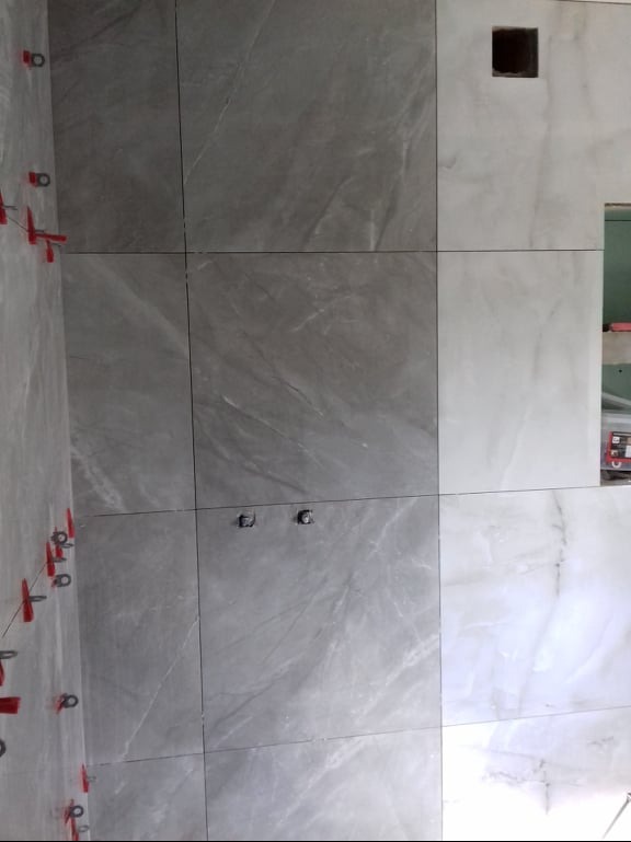 Ściana w trakcie układania płytek imitujących marmur, z widocznymi czerwonymi klipsami poziomującymi, dwa otwory na instalację elektryczną i otwór wentylacyjny.