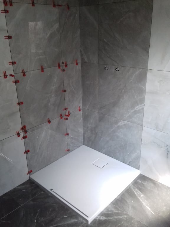 Szare płytki imitujące marmur w narożnej kabinie prysznicowej, z widocznymi czerwonymi krzyżykami dystansowymi i białym brodzikiem z odpływem.