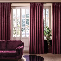 PPHU ROLETY24 ROBERT JAROSŁAWSKI - Elegancki salon z bordowymi zasłonami na szynach, dopasowaną sofą z aksamitu i nowoczesnymi dekoracjami ściennymi.
