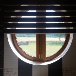 PPHU ROLETY24 ROBERT JAROSŁAWSKI - Okrągłe okno z drewnianą ramą i zamontowaną roletą dzień noc z widokiem na wiejski krajobraz z polem i ogrodzeniem.