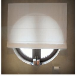 PPHU ROLETY24 ROBERT JAROSŁAWSKI - Biała, zwinięta roleta materiałowa w oknie o okrągłym kształcie. Na parapecie okna stoją dwie świeczki na małej srebrnej tacy.