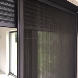 PPHU ROLETY24 ROBERT JAROSŁAWSKI - Zamknięta moskitiera przesuwna w ciemnej ramie, obok częściowo zwiniętej rolety zewnętrznej, widok z wnętrza pomieszczenia.