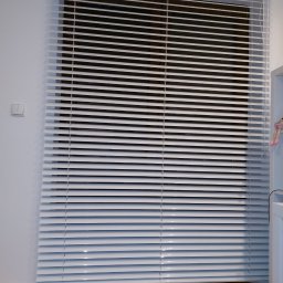 PPHU ROLETY24 ROBERT JAROSŁAWSKI - Zamknięte białe żaluzje aluminiowe na oknie, widoczny fragment ściany z włącznikiem i drewniana podłoga.