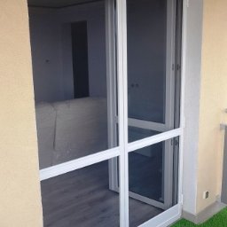 PPHU ROLETY24 ROBERT JAROSŁAWSKI - Biała moskitiera ramkowa zamontowana w drzwiach balkonowych, widok z zewnątrz na sztuczną trawę na balkonie i wnętrze pomieszczenia.