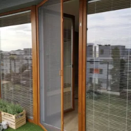 Otwarta moskitiera ramkowa w drewnianej ramie na balkonie z sztuczną trawą i roletami wewnętrznymi, widok na budynki za oknem.