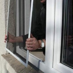 PPHU ROLETY24 ROBERT JAROSŁAWSKI - Montaż moskitiery ramkowej w oknie PCV, widoczne dłonie montera i zegarek na ręce.