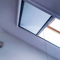 PPHU ROLETY24 ROBERT JAROSŁAWSKI - Zamknięta, jasna plisa okienna zamontowana na oknie dachowym, widoczna z dołu pod kątem, z białym mechanizmem zwijającym i cienką linką sterującą.