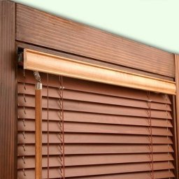 PPHU ROLETY24 ROBERT JAROSŁAWSKI - Brązowe żaluzje drewniane z widocznym mechanizmem sznurkowym i drewnianą ramą okienną w ciepłym oświetleniu.