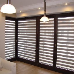 PPHU ROLETY24 ROBERT JAROSŁAWSKI - Eleganckie rolety dzień i noc w brązowych ramach okiennych, widoczne z wnętrza pokoju z białym fotelem i drewnianą podłogą, oświetlone lampami sufitowymi.