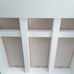 PPHU ROLETY24 ROBERT JAROSŁAWSKI - Beżowe plisy zamontowane w oknach dachowych, widok z dołu, białe belki konstrukcyjne sufitu, realizacja w Chmielnie.