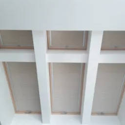 Beżowe plisy zamontowane w oknach dachowych, widok z dołu, białe belki konstrukcyjne sufitu, realizacja w Chmielnie.