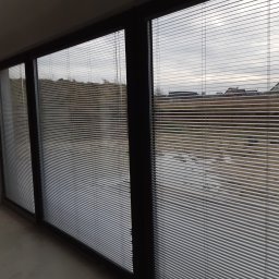 PPHU ROLETY24 ROBERT JAROSŁAWSKI - Zamknięte, poziome, aluminiowe żaluzje w oknach przesuwnych, widok na zewnątrz z szarą, pochmurną pogodą.