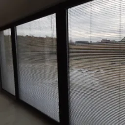 Zamknięte, poziome, aluminiowe żaluzje w oknach przesuwnych, widok na zewnątrz z szarą, pochmurną pogodą.