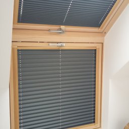 PPHU ROLETY24 ROBERT JAROSŁAWSKI - Szare, harmonijkowe osłony okienne zamontowane w drewnianej ramie okna dachowego, widoczne w dwóch częściach: górnej i dolnej.