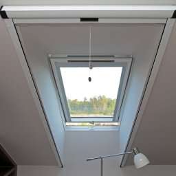 PPHU ROLETY24 ROBERT JAROSŁAWSKI - Okno dachowe z częściowo zwiniętą roletą materiałową w kasecie, widok na zewnątrz z zielenią, sznurek do regulacji zwisa pionowo.