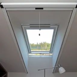 Okno dachowe z częściowo zwiniętą roletą materiałową w kasecie, widok na zewnątrz z zielenią, sznurek do regulacji zwisa pionowo.