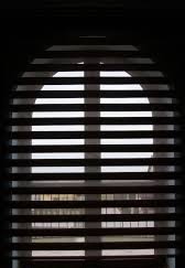 Zasłonięte okno z widocznymi poziomymi lamelami, tworzącymi efekt światłocienia. Silne oświetlenie z zewnątrz przez okrągłe okno.