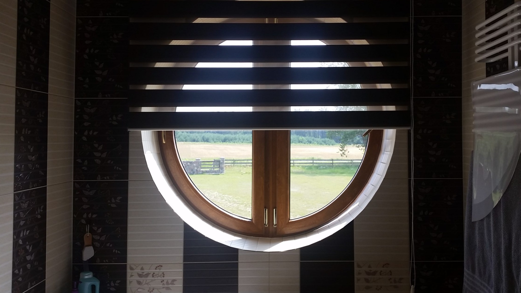 Okrągłe okno z drewnianą ramą i zamontowaną roletą dzień noc z widokiem na wiejski krajobraz z polem i ogrodzeniem.