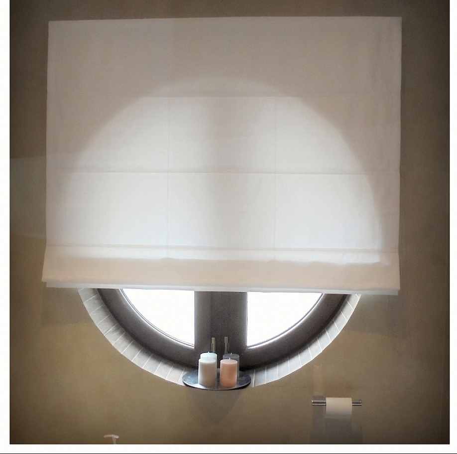 Biała, zwinięta roleta materiałowa w oknie o okrągłym kształcie. Na parapecie okna stoją dwie świeczki na małej srebrnej tacy.