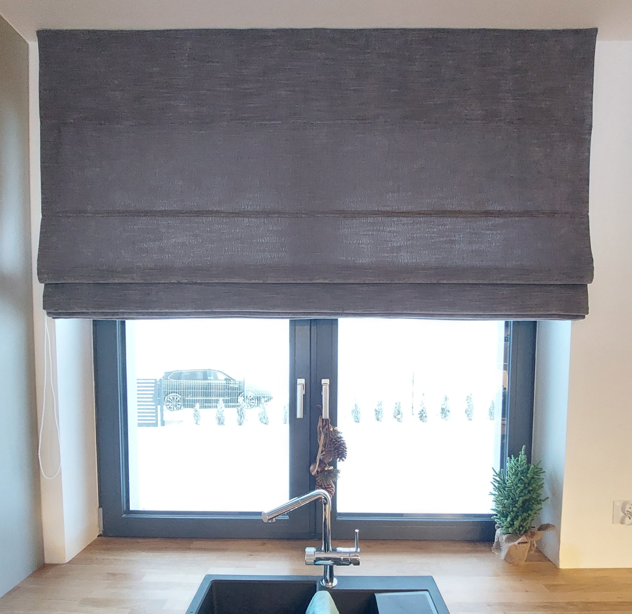 Szara roleta rzymska zamontowana w kuchni nad oknem z widokiem na zasypany śniegiem ogród i zaparkowany samochód.