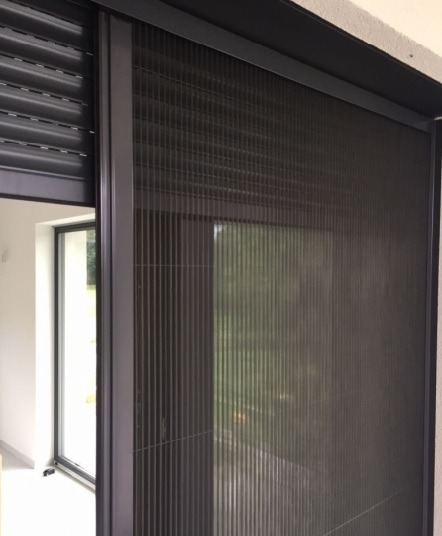 Zamknięta moskitiera przesuwna w ciemnej ramie, obok częściowo zwiniętej rolety zewnętrznej, widok z wnętrza pomieszczenia.