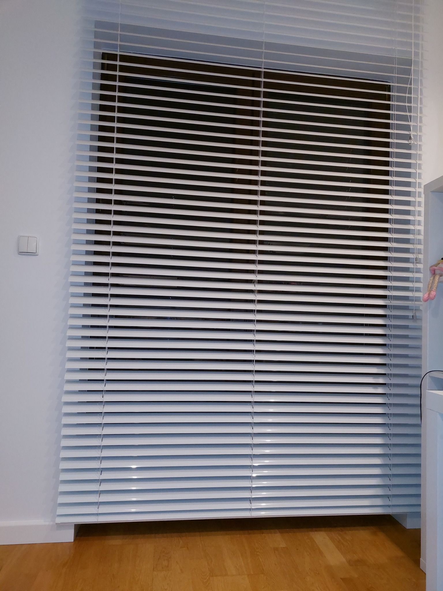 Zamknięte białe żaluzje aluminiowe na oknie, widoczny fragment ściany z włącznikiem i drewniana podłoga.