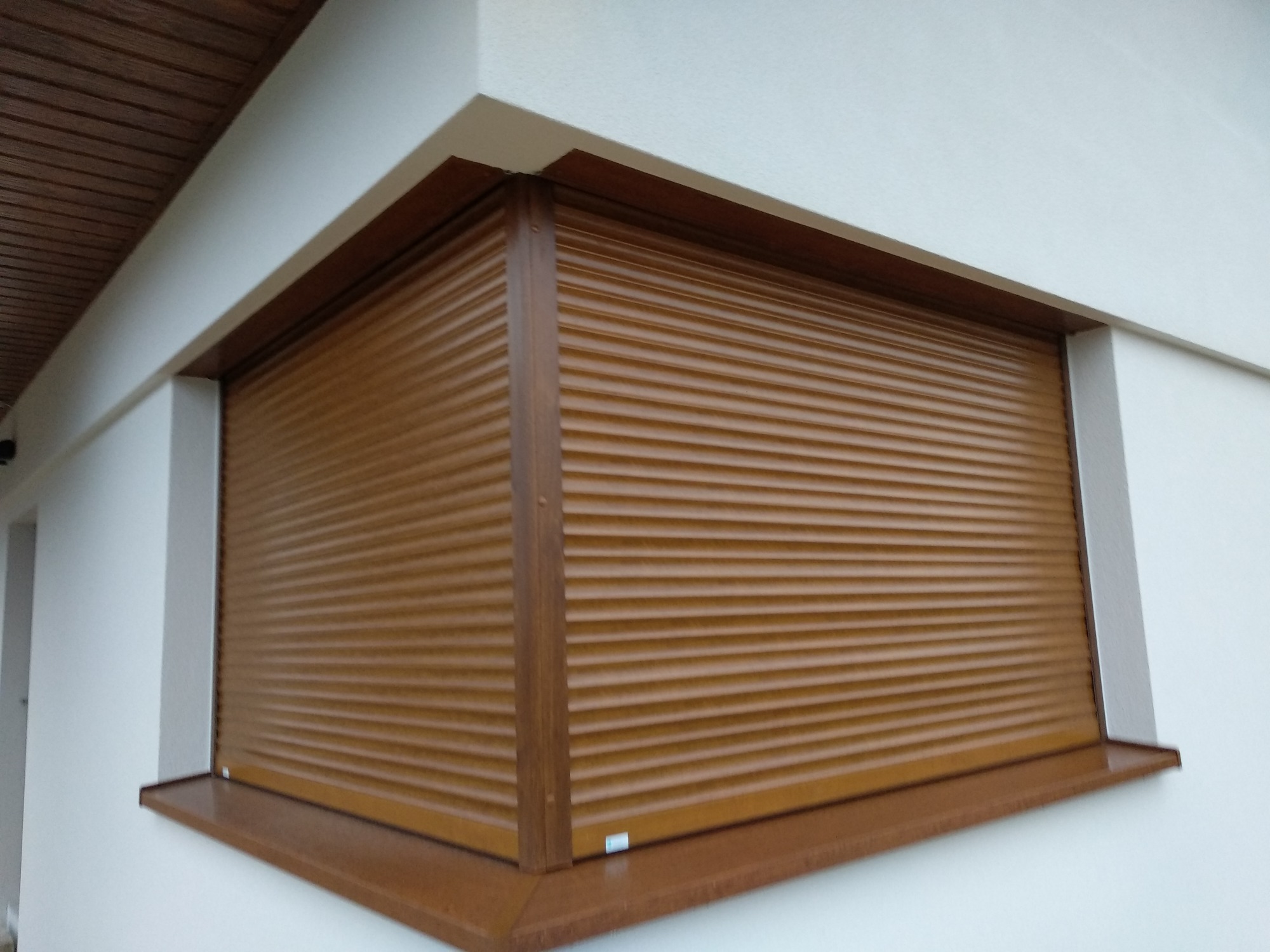 Zewnętrzna brązowa roleta zwinięta na oknie budynku z białymi ścianami, widoczne drewniane wykończenia ramy okiennej i parapetu.