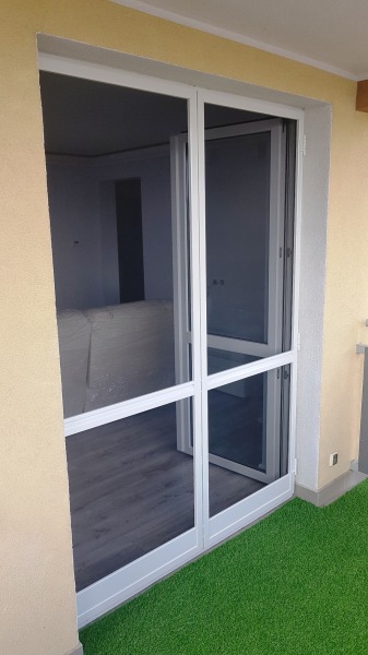 Biała moskitiera ramkowa zamontowana w drzwiach balkonowych, widok z zewnątrz na sztuczną trawę na balkonie i wnętrze pomieszczenia.